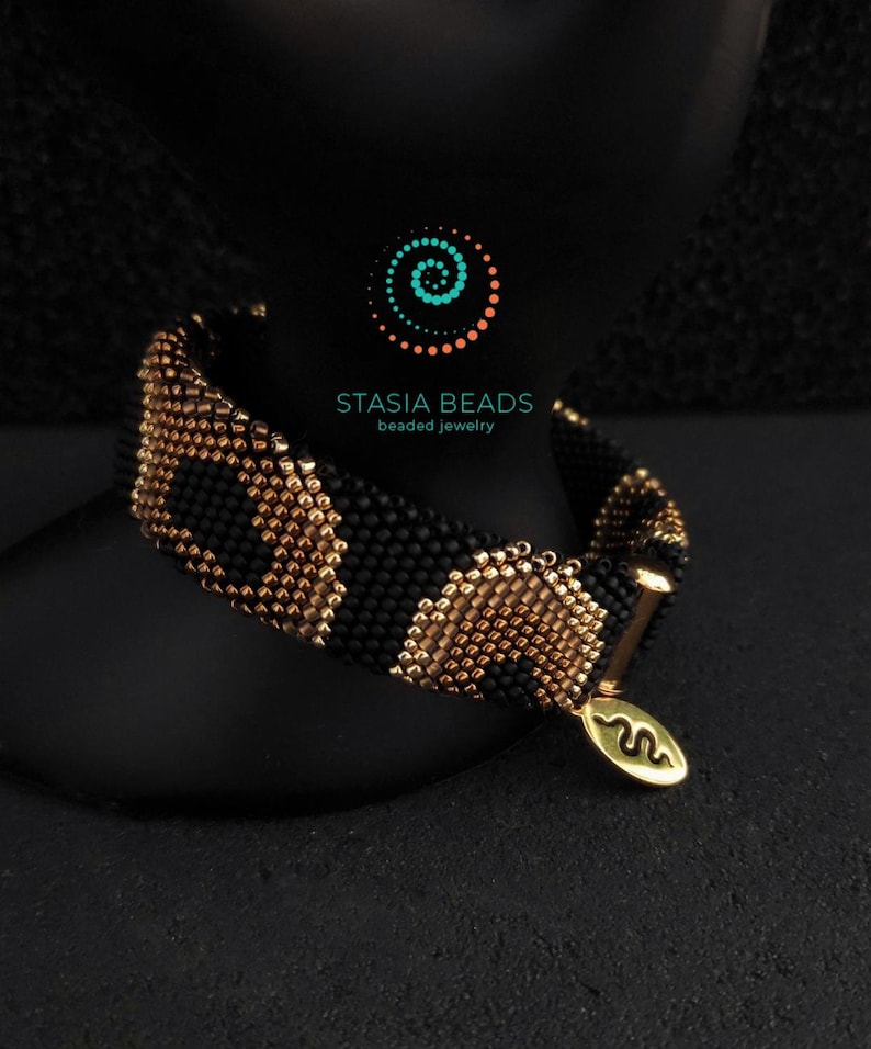 May include: A black and gold beaded bracelet with a snake pattern. The bracelet is made of small, round beads and has a gold-coloured clasp and charm. The charm has a snake symbol and the text "STASIA BEADS beaded jewelry".