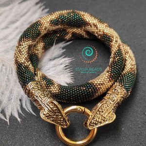 May include: A beaded necklace with a snake design in shades of green and gold. The necklace has two gold snake head clasps and a gold lobster clasp.