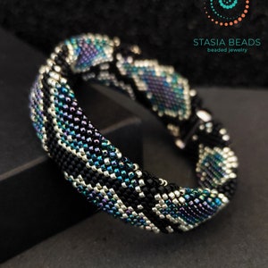 Puede incluir: Un brazalete de cuentas negras, plateadas y azules con un patrón geométrico. El brazalete está hecho con cuentas pequeñas e iridiscentes y es una pieza de joyería llamativa.
