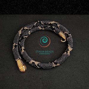 May include: A black and gold beaded snake necklace with a gold snake head clasp. The necklace is made of small, round beads and has a geometric pattern.