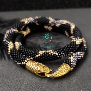 May include: A coiled beaded necklace featuring a black, gold, and iridescent purple pattern. The necklace ends in two gold-toned metal snake head clasps that hook together. The text STASIA BEADS beaded jewelry is printed in the center of the coil.