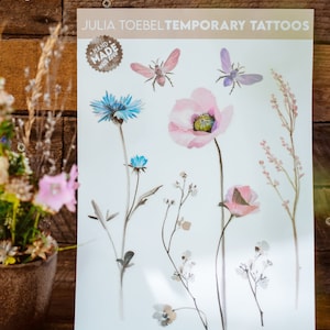 以下が含まれることがあります： 水彩画で描かれた花と昆虫のイラストが特徴の、一時的なタトゥーのシート。ピンクのポピー、青いヤグルマギク、繊細な小枝のデザインが含まれています。「JULIA TOEBEL TEMPORARY TATTOOS」の文字が上部に印刷され、「HAND MADE with love」のシールが付いています。