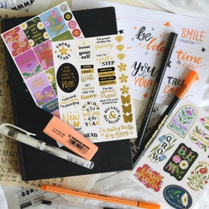 Op de afbeelding: Een verzameling gouden en zwarte motiverende stickers met zinnen als "Investeer in jezelf", "De beste tijd om te beginnen is nu", "Ik creëer het leven van mijn dromen" en "Bloei elke dag". De stickers zitten op een zwart notitieboek met een roze markeerstift en oranje en zwarte stiften.