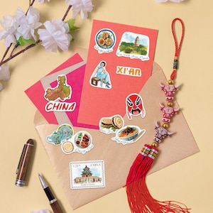 May include: A set of colorful stickers featuring images of Chinese landmarks, food, and culture. The stickers are arranged on a red and brown envelope with a red tassel hanging from the top. The stickers include a map of China, a pagoda, a traditional Chinese mask, a bowl of noodles, and a teapot.