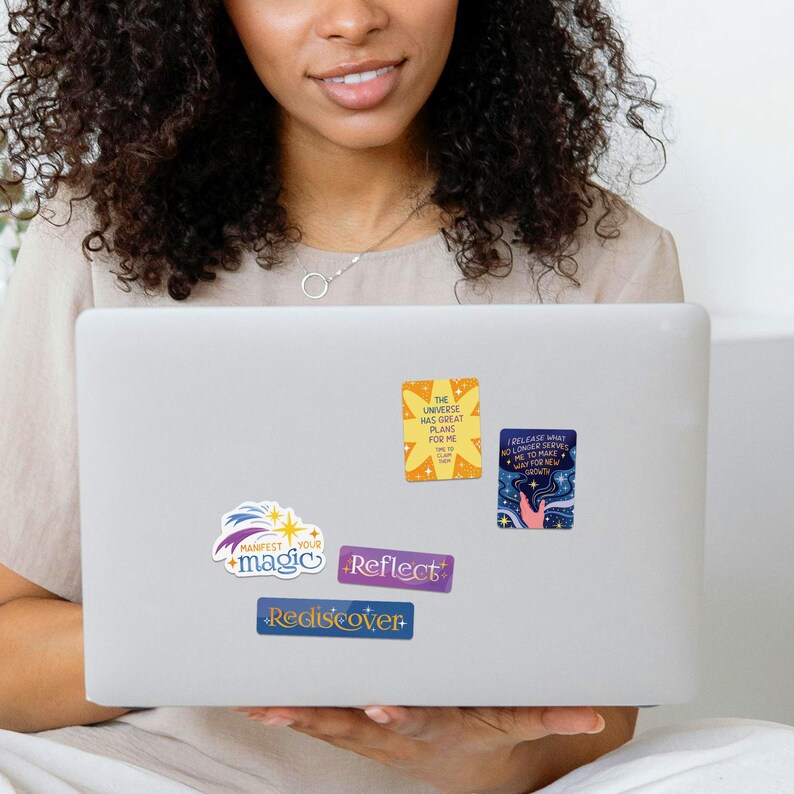 Może przedstawiać: Laptop z pięcioma kolorowymi naklejkami na tylnej stronie. Naklejki zawierają inspirujące wiadomości, w tym "Wszechświat ma dla mnie wielkie plany", "Zwalniam to, co mi już nie służy, aby zrobić miejsce na nowy wzrost", "Manifestuj swoją magię", "Reflektuj" i "Odkryj na nowo".