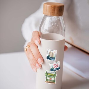 Puede incluir: Una botella de agua reutilizable blanca con una tapa de madera y tres pegatinas con tem&aacute;tica parisina. Las pegatinas presentan ilustraciones de la Torre Eiffel, un caf&eacute; parisino y el texto "Au Revoir".