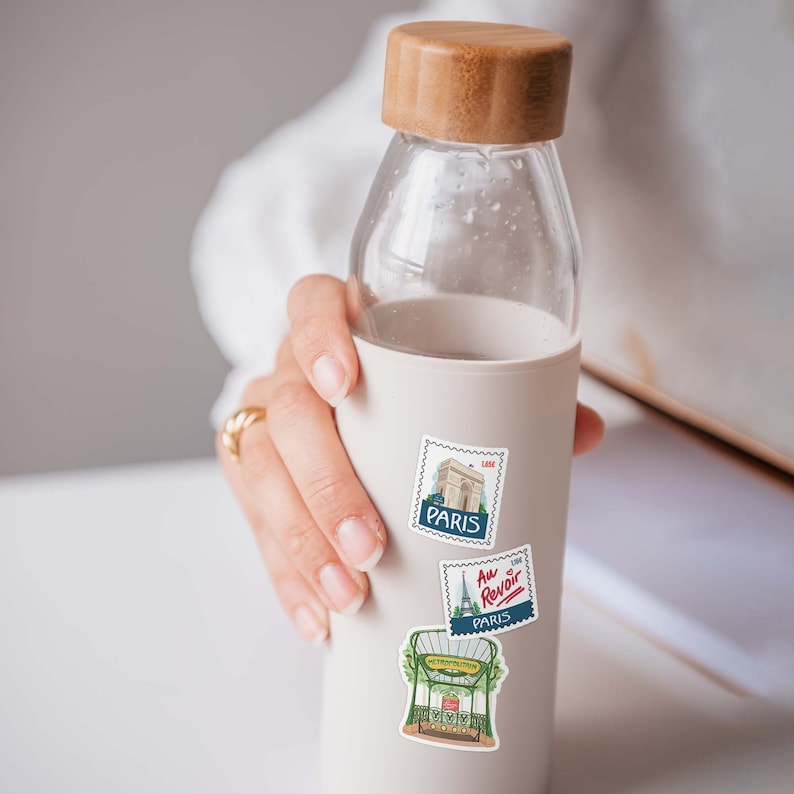 Puede incluir: Una botella de agua reutilizable blanca con una tapa de madera y tres pegatinas con tem&aacute;tica parisina. Las pegatinas presentan ilustraciones de la Torre Eiffel, un caf&eacute; parisino y el texto "Au Revoir".
