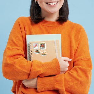 May include: A person holding a brown notebook with a spiral binding. The notebook has several colorful stickers on the front, including a red building, a bowl of noodles, and a flower.