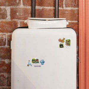 May include: White suitcase with colorful travel stickers featuring images of the Great Wall of China, a panda bear, a blue and white porcelain vase, and a building in Shanghai, China.