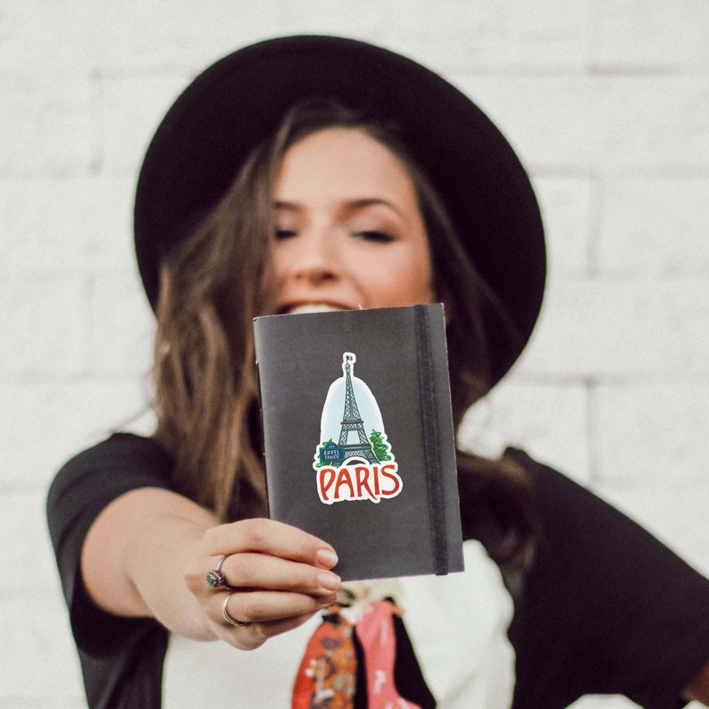 Puede incluir: Una mujer con un sombrero negro y una camisa negra sostiene un cuaderno negro con una pegatina de la Torre Eiffel y la palabra "Par&iacute;s" en &eacute;l.