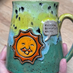May include: Handmade ceramic mug with a smiling sun design in orange and black. The mug has a green, yellow, and blue glaze with the words "FREEDOM, DEMOCRACY, TRUTH" in a speech bubble. The mug has a handle.