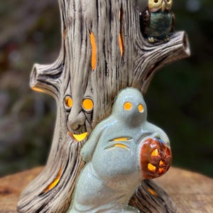 May include: A ceramic Halloween decoration featuring a tree trunk with a carved face and glowing orange lights. A ghost figure holds a carved pumpkin, and an owl sits on a branch. The color palette includes beige, green, brown, and orange.