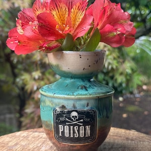 May include: A handmade ceramic vase with a turquoise and brown glaze, featuring a "POISON" label with a skull and crossbones. The vase holds vibrant red and yellow flowers, creating a striking contrast. The vase is approximately 5 inches tall.