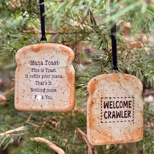 May include: Two tan, speckled, toast-shaped ornaments hanging from black ribbons. The left ornament reads: Mana Toast. This is Toast. It refills your mana. That’s it. Nothing more. F*ck You. The right ornament reads: WELCOME CRAWLER inside a dashed box.