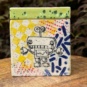 May include: A ceramic box with a green lid and a colorful robot design. The box has a white background with yellow, red, and blue geometric shapes.