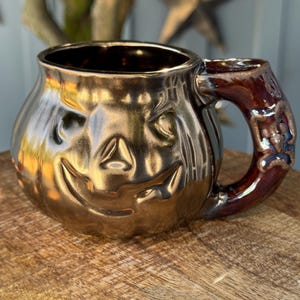 May include: A metallic gold ceramic mug shaped like a jack-o'-lantern with a carved face. The handle is a dark brown with a decorative design. The mug is sitting on a wooden surface.