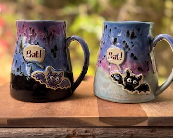 May include: Two ceramic mugs with a Halloween theme. The mugs are blue, purple, and black with a bat design and the word "Bat!" in a speech bubble. The mugs are on a wooden surface.