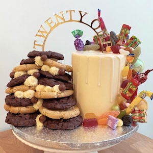 May include: A white cake with a gold 'Twenty' topper and a drip of white icing. The cake is decorated with a variety of colourful sweets, including gummy sweets, chocolate sweets, and lollipops. The cake is surrounded by a stack of chocolate and vanilla biscuits.