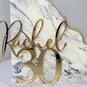 Könnte beinhalten: Goldfarbener, spiegelnder Acryl-Kuchentopper mit dem Namen "Rachel" und der Zahl "30" in einer kursiv geschriebenen Schrift.