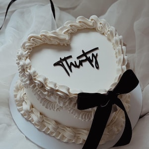 Puede incluir: Un pastel blanco en forma de corazón con glaseado blanco y una cinta negra. El pastel está decorado con la palabra "Thirty" en glaseado negro.