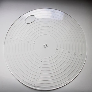 Peut inclure: Une règle circulaire en plastique transparent avec des cercles concentriques et des chiffres de 1 à 12. La règle a un petit trou au centre et un signe plus au milieu.
