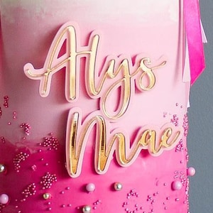 Könnte beinhalten: Rosa und goldener Tortendekor mit dem Namen "Alys Mae" in Kursivschrift.