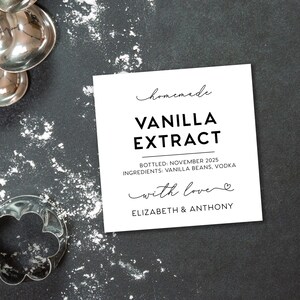 May include: A square white label with black text reading Homemade VANILLA EXTRACT, BOTTLED: NOVEMBER 2025, INGREDIENTS: VANILLA BEANS, VODKA, with love, ELIZABETH & ANTHONY, surrounded by silver measuring spoons, a metal cup, a sieve, and a flower cutter.