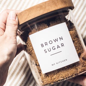 May include: A clear glass storage jar with a wooden lid filled with brown sugar. A white square label on the front features the black text BROWN SUGAR with a horizontal line underneath and the words MY KITCHEN printed below the line.