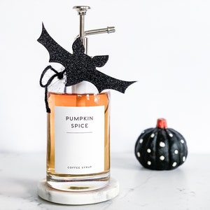 Pu&ograve; includere: Una bottiglia di vetro con un erogatore a pompa riempita di liquido arancione. La bottiglia ha un'etichetta bianca su cui &egrave; scritto "Pumpkin Spice Coffee Syrup". Un ritaglio di pipistrello nero &egrave; attaccato all'erogatore a pompa.