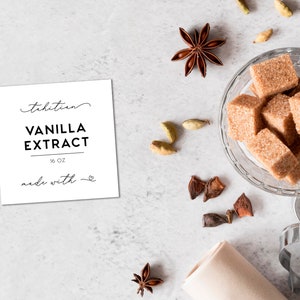 May include: A square white label with black text reading Tahitian VANILLA EXTRACT 16 OZ made with a heart symbol. Nearby are brown sugar cubes in a glass bowl, star anise, cardamom pods, and a metal heart-shaped cookie cutter on a light surface.