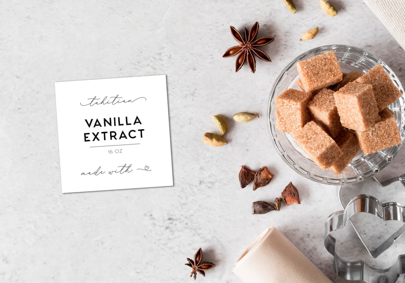 May include: A square white label with black text reading Tahitian VANILLA EXTRACT 16 OZ made with a heart symbol. Nearby are brown sugar cubes in a glass bowl, star anise, cardamom pods, and a metal heart-shaped cookie cutter on a light surface.