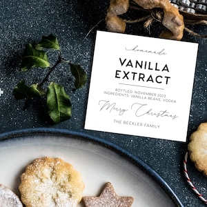 May include: A square white label with black text reading homemade VANILLA EXTRACT, BOTTLED: NOVEMBER 2023, INGREDIENTS: VANILLA BEANS, VODKA, Merry Christmas, and THE BECKLER FAMILY, surrounded by white snowflake cutouts and cookies.