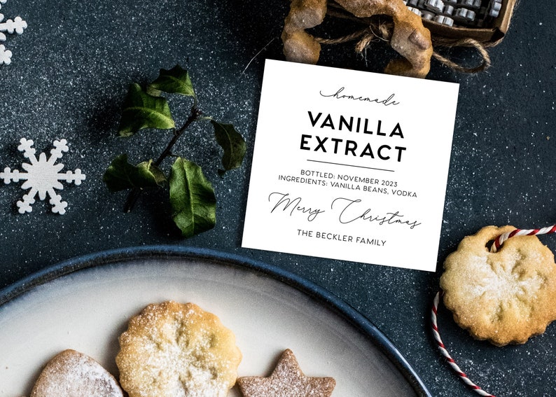 May include: A square white label with black text reading homemade VANILLA EXTRACT, BOTTLED: NOVEMBER 2023, INGREDIENTS: VANILLA BEANS, VODKA, Merry Christmas, and THE BECKLER FAMILY, surrounded by white snowflake cutouts and cookies.