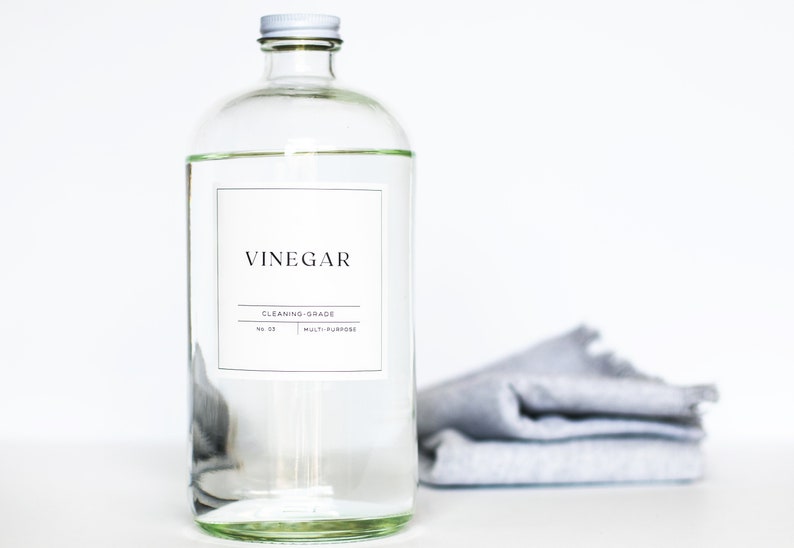 May include: A clear glass bottle with a white label that reads "Vinegar Cleaning Grade No. 01 Multi-Purpose". The bottle is sitting on a white surface with a gray cloth folded in the background.