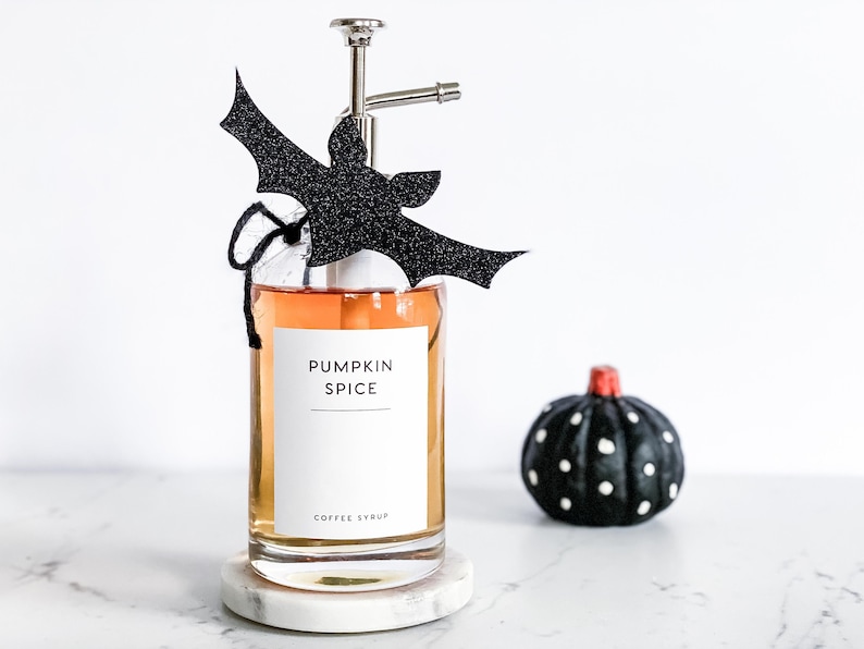Pu&ograve; includere: Una bottiglia di vetro con un erogatore a pompa riempita di liquido arancione. La bottiglia ha un'etichetta bianca su cui &egrave; scritto "Pumpkin Spice Coffee Syrup". Un ritaglio di pipistrello nero &egrave; attaccato all'erogatore a pompa.