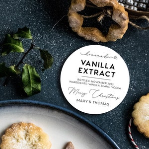 May include: A round white label with black text reading homemade VANILLA EXTRACT, BOTTLED: NOVEMBER 2023, INGREDIENTS: VANILLA BEANS, VODKA, Merry Christmas, and MARY & THOMAS, surrounded by holiday cookies, a green sprig, and a snowflake cutout.