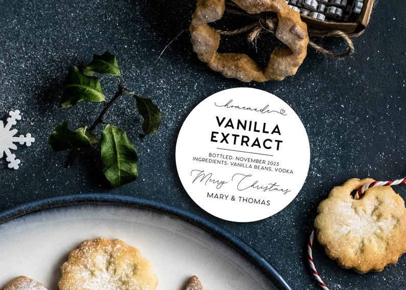 May include: A round white label with black text reading homemade VANILLA EXTRACT, BOTTLED: NOVEMBER 2023, INGREDIENTS: VANILLA BEANS, VODKA, Merry Christmas, and MARY & THOMAS, surrounded by holiday cookies, a green sprig, and a snowflake cutout.
