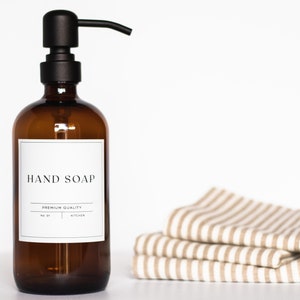 May include: Amber glass bottle with a black pump dispenser and a white label that reads "HAND SOAP" and "PREMIUM QUALITY". The bottle is sitting on a white surface next to a stack of striped fabric towels.