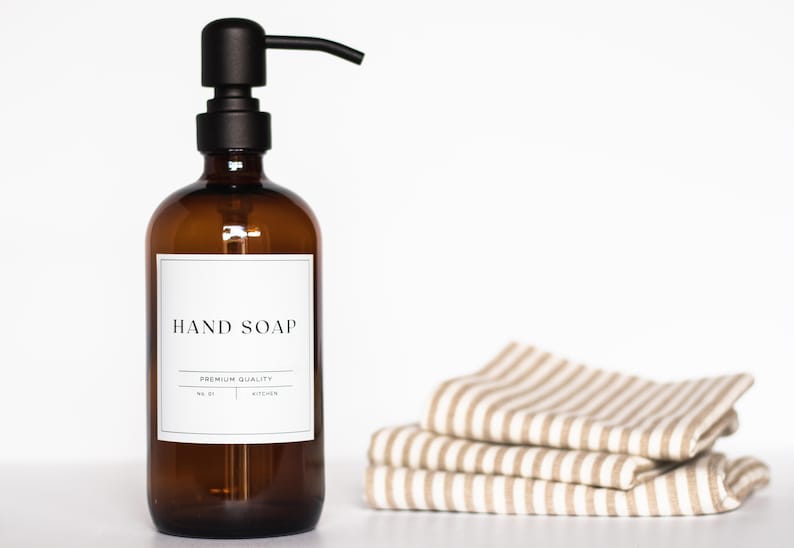 May include: Amber glass bottle with a black pump dispenser and a white label that reads "HAND SOAP" and "PREMIUM QUALITY". The bottle is sitting on a white surface next to a stack of striped fabric towels.