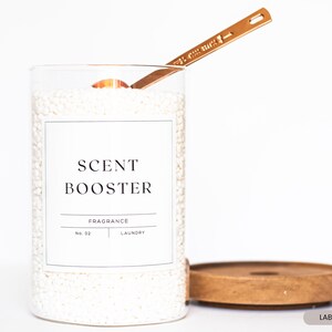May include: A clear glass jar filled with white laundry scent booster beads. The jar has a white label with black text that reads "SCENT BOOSTER", "FRAGRANCE", "NO. 02", and "LAUNDRY". A gold metal spoon is resting on the top of the jar.