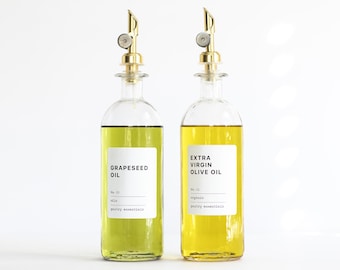 May include: Two clear glass bottles with gold-colored metal pour spouts. The left bottle contains light green grapeseed oil with a white label reading GRAPESEED OIL No. 05 oils pantry essentials. The right bottle contains yellow extra virgin olive oil.