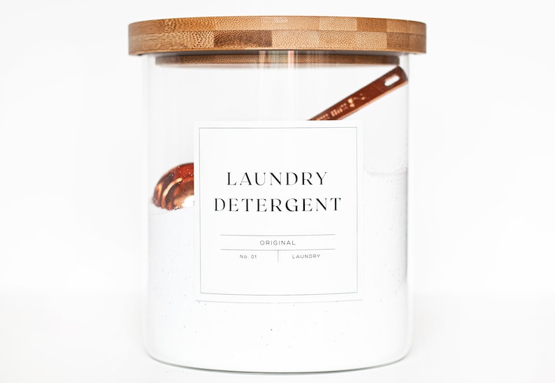 May include: A glass jar with a bamboo lid filled with white laundry detergent powder. A small copper scoop is inside the jar. A white label on the jar reads "LAUNDRY DETERGENT ORIGINAL NO. 01 LAUNDRY"