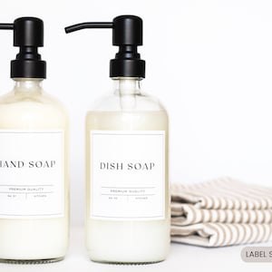 May include: Two clear glass bottles with black pump tops, filled with liquid soap. The bottles have white labels with black text that reads "HAND SOAP" and "DISH SOAP". The labels also include the text "PREMIUM QUALITY" and "KITCHEN". The bottles are sitting on a white surface with a stack of striped towels in the background. The text "LABEL SIZE - 3" x 3" is printed below the towels.