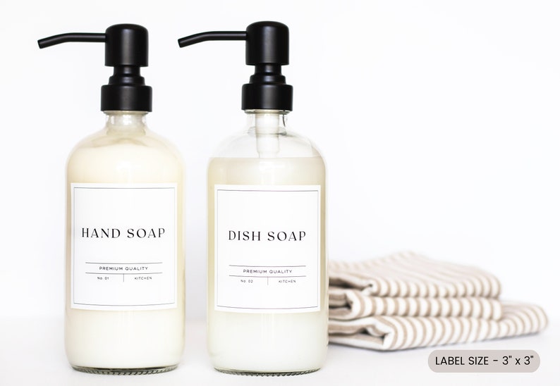 May include: Two clear glass bottles with black pump tops, filled with liquid soap. The bottles have white labels with black text that reads "HAND SOAP" and "DISH SOAP". The labels also include the text "PREMIUM QUALITY" and "KITCHEN". The bottles are sitting on a white surface with a stack of striped towels in the background. The text "LABEL SIZE - 3" x 3" is printed below the towels.