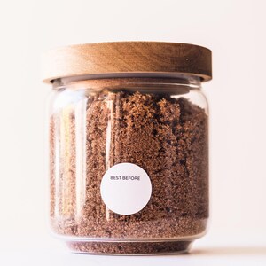 May include: A glass jar with a wooden lid filled with brown sugar. A white sticker on the jar reads "BEST BEFORE".