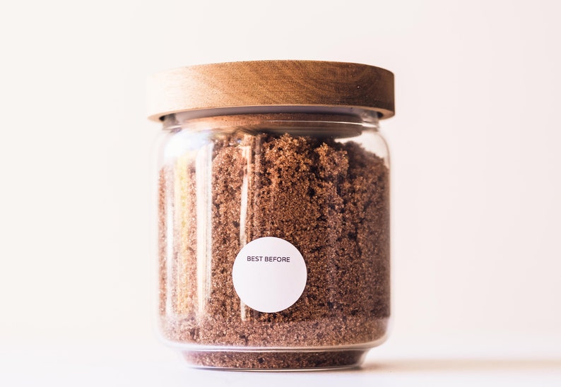 May include: A glass jar with a wooden lid filled with brown sugar. A white sticker on the jar reads "BEST BEFORE".