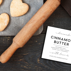 May include: A wooden rolling pin, heart-shaped cookies on a gray board, and a white square label that reads: homemade CINNAMON BUTTER DECEMBER 2023 INGREDIENTS: HEAVY CREAM, SUGAR, SALT, CINNAMON with love EVELYN HARDCASTLE.