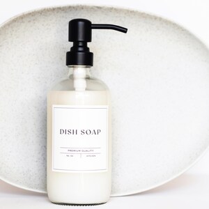 May include: A clear glass bottle with a black pump dispenser filled with white dish soap. The bottle has a white label with the text "DISH SOAP" and "PREMIUM QUALITY" in black lettering.