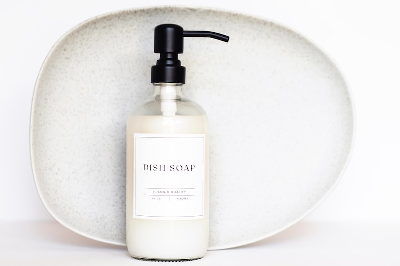 May include: A clear glass bottle with a black pump dispenser filled with white dish soap. The bottle has a white label with the text "DISH SOAP" and "PREMIUM QUALITY" in black lettering.