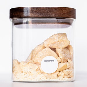 May include: A glass jar with a wooden lid filled with chunks of light brown shea butter. A white label on the jar reads "BEST BEFORE".